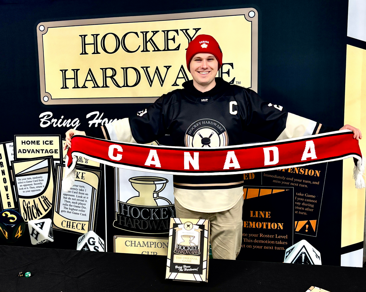 Hockey Hardware creator Billy Morrison wearing a Hockey Hardware jersey and a Canada World Juniors hat, standing in front of a Hockey Hardware backdrop wall holding up a red scarf that reads, 'CANADA'.