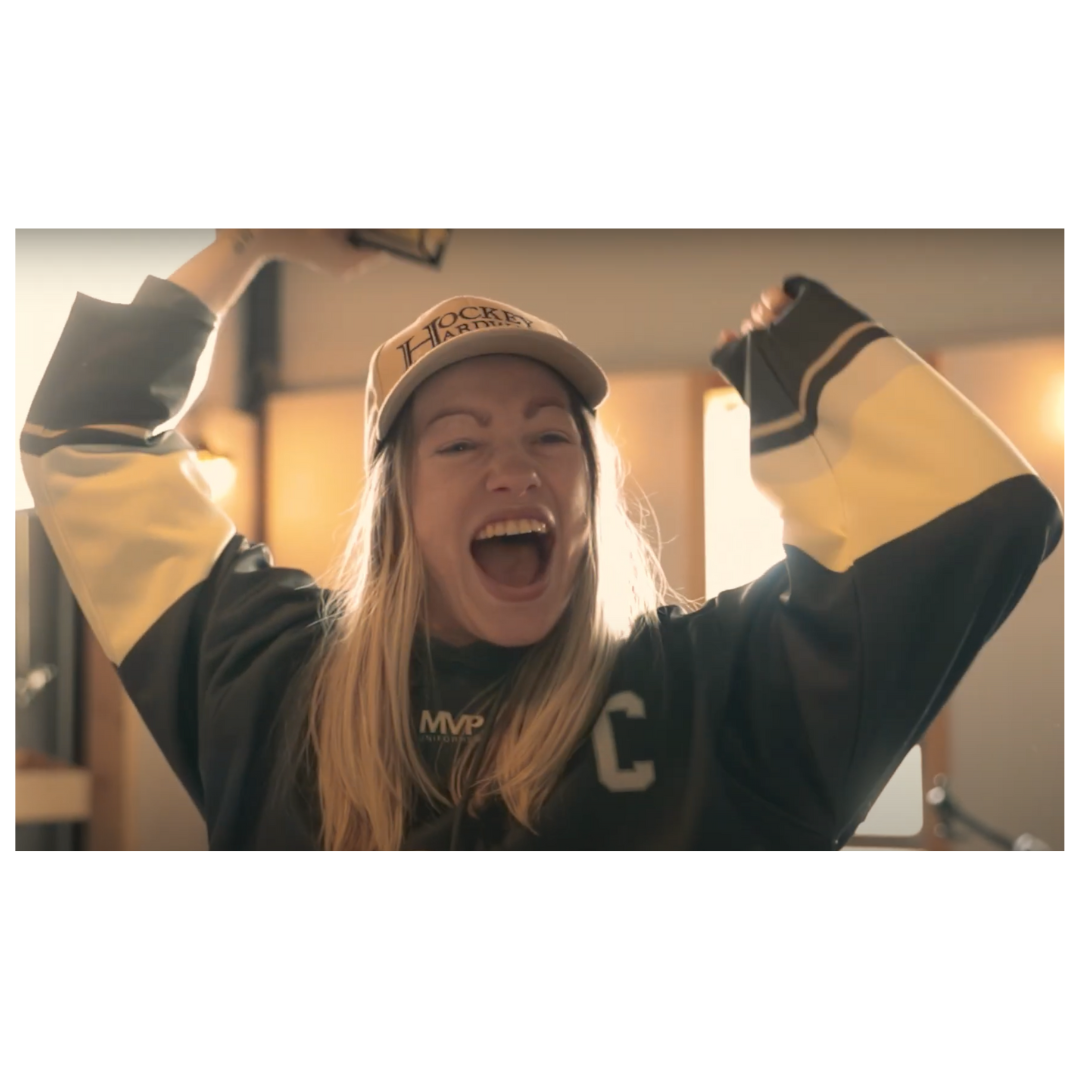 A young adult woman celebrates in her Hockey Hardware jersey and hat.