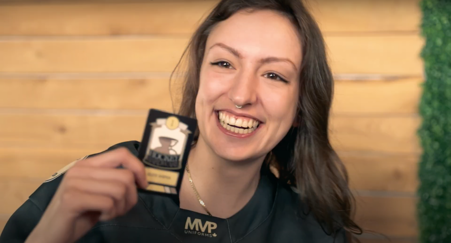 A young adult woman smiles big as she holds up a Hockey Hardware Trophy Card. 